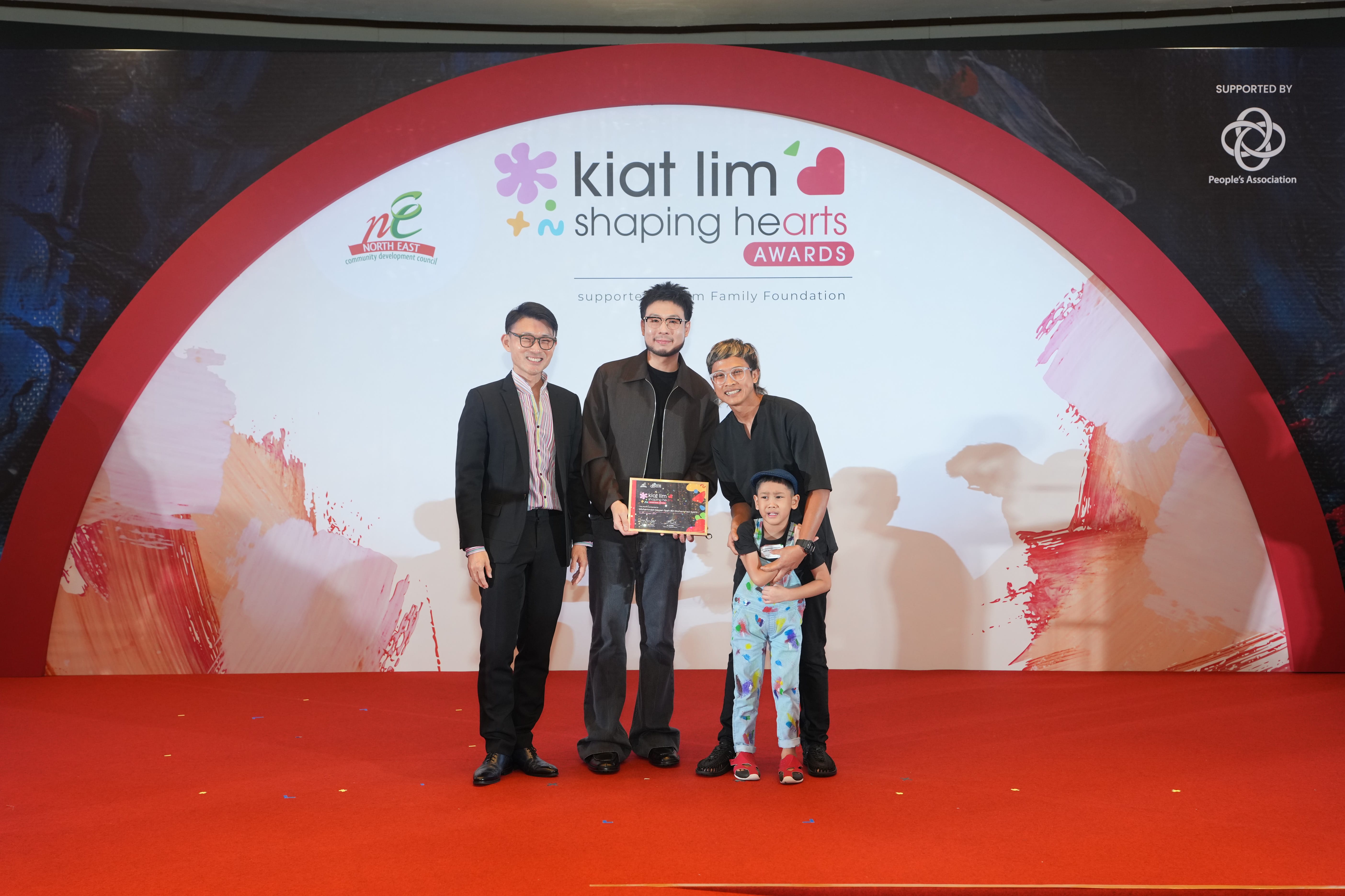 Award recipient standing on stage with the presenter, holding their certificate and posing for a group photo during the award ceremony, with a backdrop reading ‘Kiat Lim-Shaping Hearts Awards'.